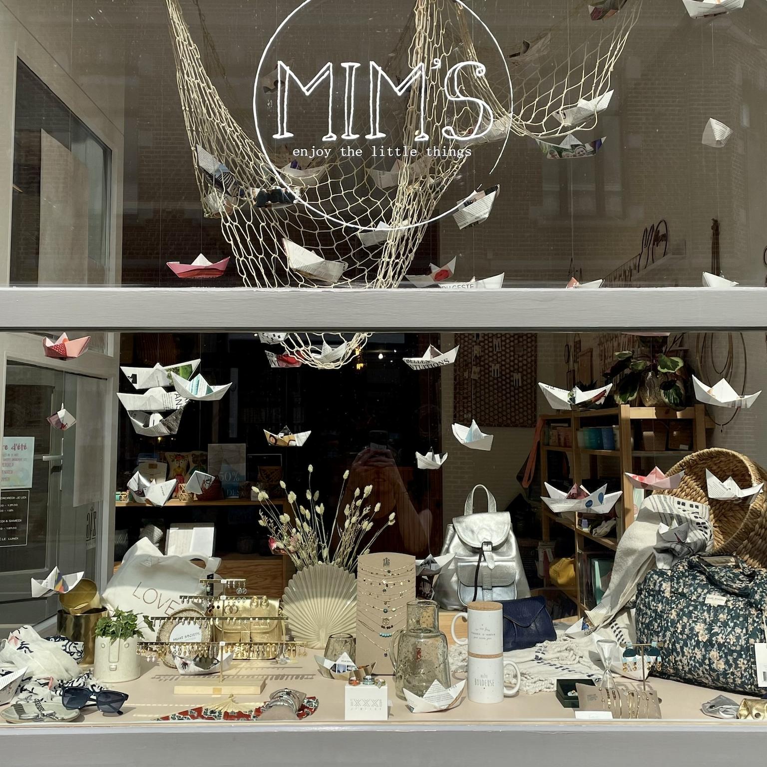 Vitrine de la boutique Mim's à Leuze-en-Hainaut avec des dizaines de bateaux en papier suspendus, entourés de sacs, tasses et colliers fantaisie