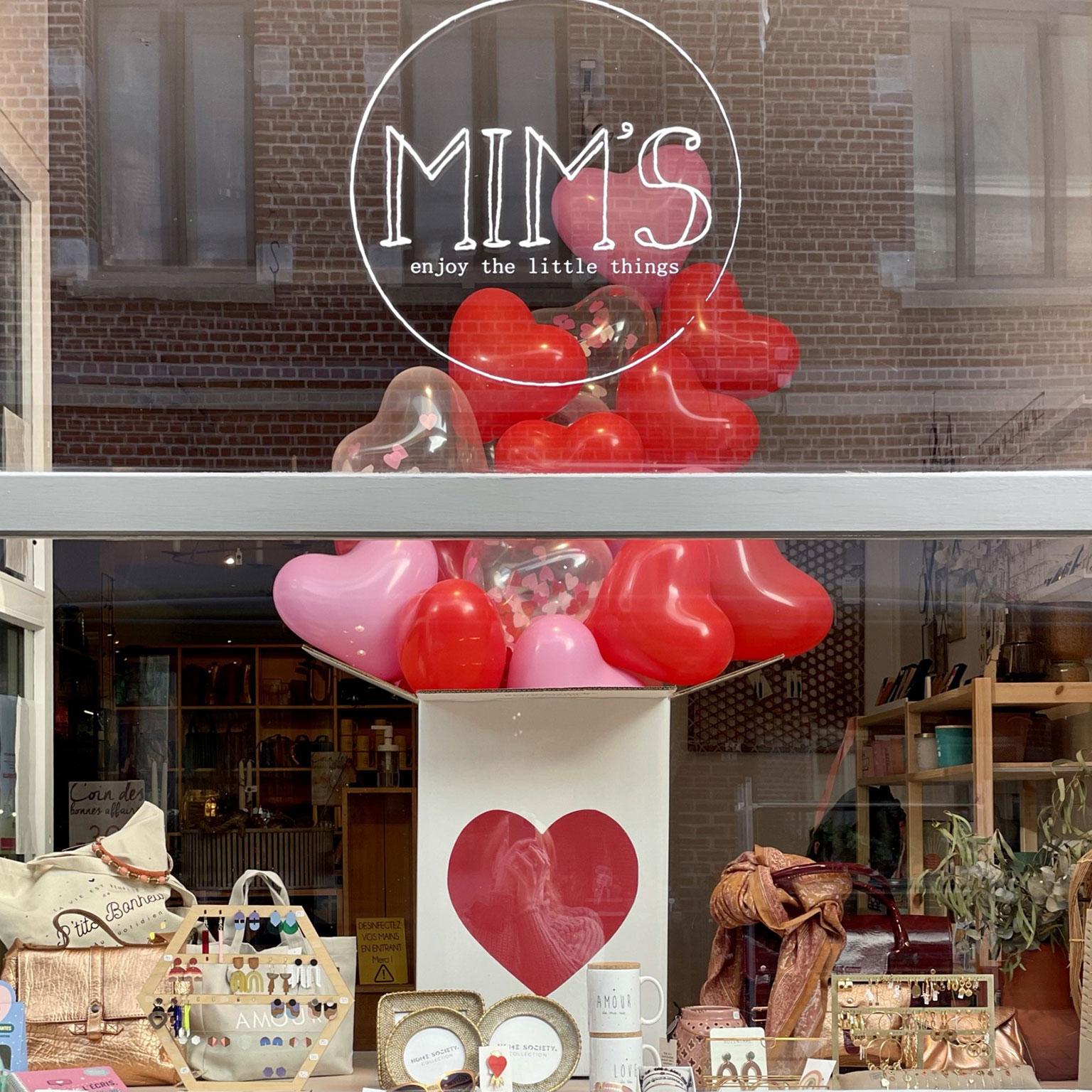 Vitrine de la boutique Mim's à Leuze sur le thème de la Saint-Valentin avec des ballons en forme de cœur, un carton décoré et des sacs, bijoux et objets déco