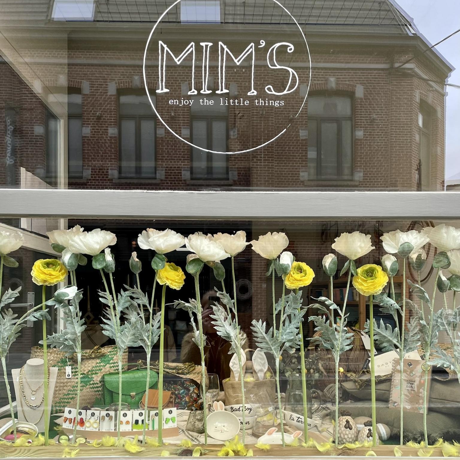 Vitrine de la boutique Mim's à Leuze-en-Hainaut décorée de fleurs blanches et jaunes, ambiance printanière avec sacs verts et bruns, bijoux, livre et objets de décoration