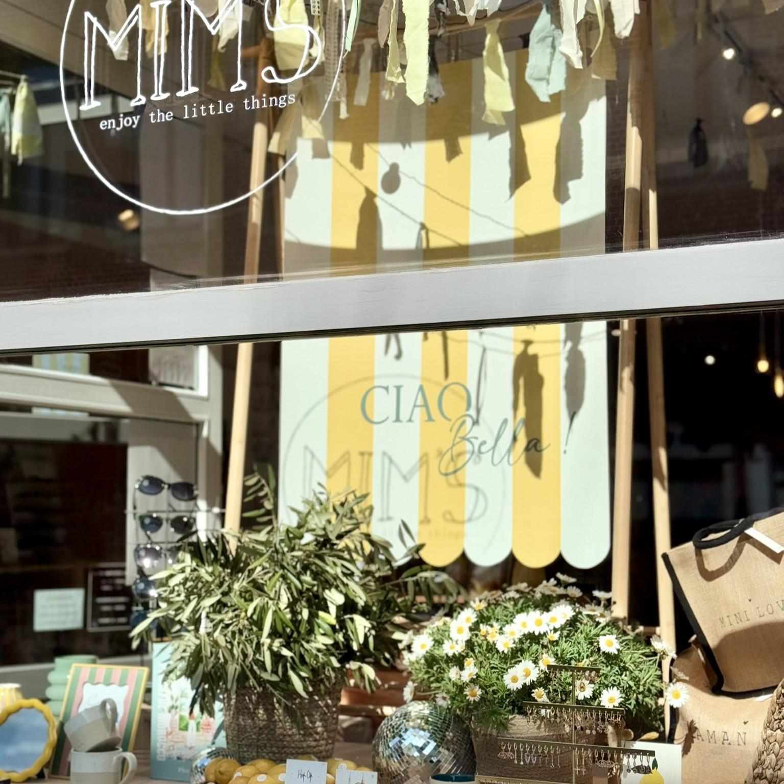 Vitrine printanière de la boutique Mim's à Leuze avec lunettes de soleil, marguerites, fleurs colorées, boule à facette et tenture rayée jaune et blanche 'Ciao Bella'