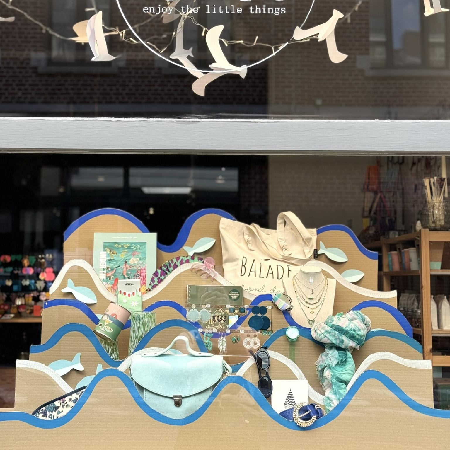 Vitrine de la boutique Mim's à Leuze-en-Hainaut décorée de vagues en bleu et beige mettant en valeur sacs, ceintures, colliers et objets de décoration